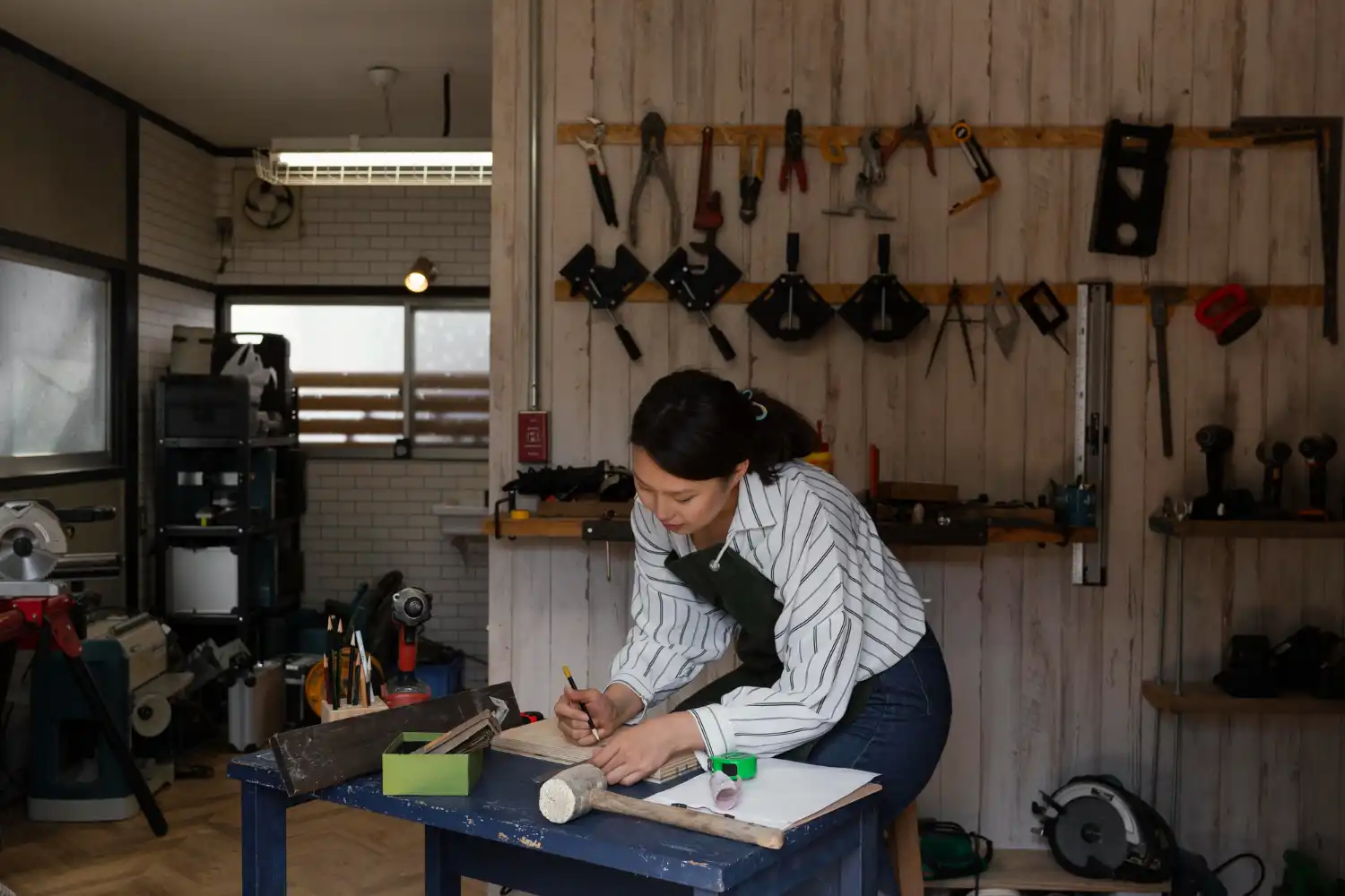 Mulher organizando itens essenciais para uma oficina eficiente, com diversas ferramentas penduradas e equipamentos de trabalho ao fundo.