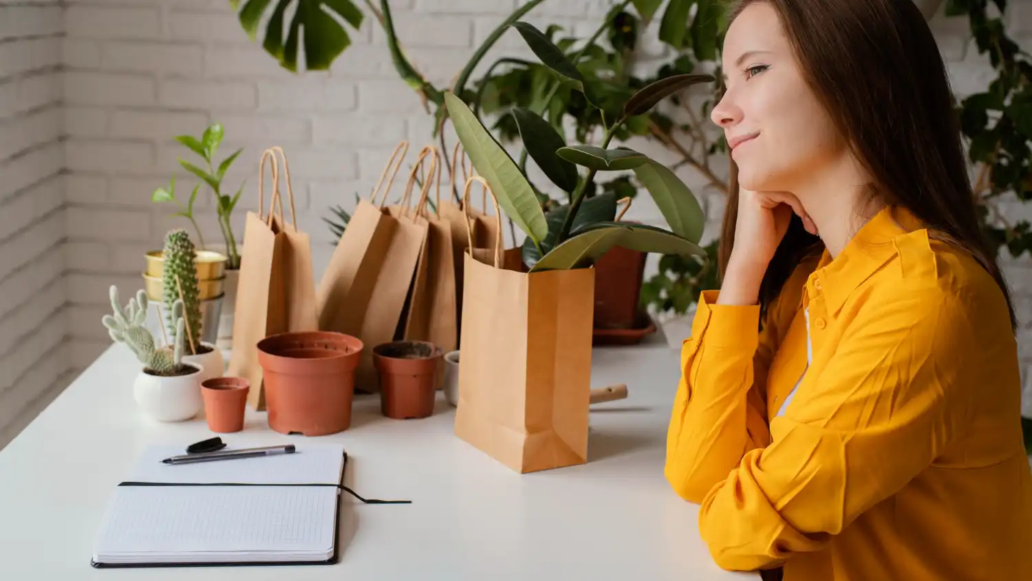 Mulher sorridente analisando presentes corporativos sustentáveis em um ambiente moderno, com diversas sacolas de papel, vasos de plantas e caderno na mesa.