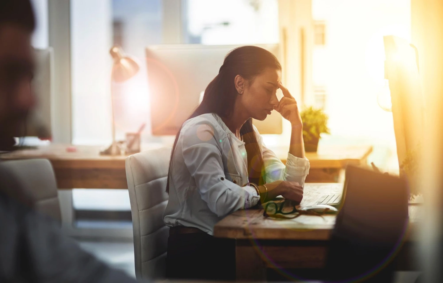 Saúde mental no trabalho: Como identificar e prevenir o esgotamento: Mulher no escritório, com postura curvada e expressão de cansaço, apoiando a cabeça com a mão enquanto trabalha em um computador ao fundo, em clima de estresse e exaustão